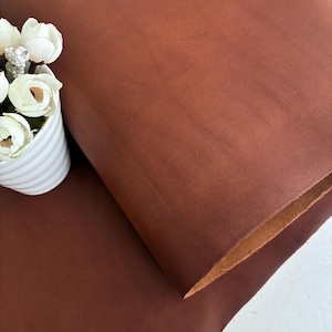 May include: A large, folded piece of smooth, matte, reddish-brown leather with a visible grain texture. Beside the leather is a white, ribbed ceramic pot containing a cluster of small, white artificial flowers with green stems and centres.