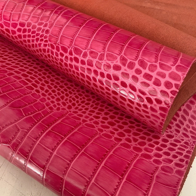 May include: Close-up of pink crocodile embossed leather. The leather has a glossy finish and a textured pattern.
