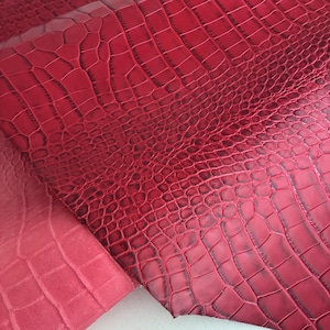 May include: A sheet of vibrant red leather featuring a prominent embossed crocodile skin texture. The surface displays a repeating pattern of raised, irregular scales with darker red shading within the crevices, creating a deep, dimensional look.