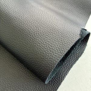 Op de afbeelding: Een stuk zwart leermateriaal met een uitgesproken korrelige textuur op het oppervlak. Het materiaal is omgevouwen om de gladde, donkere en vezelige onderkant te onthullen, wat de dikte en de gelaagde kwaliteit van de huid laat zien.
