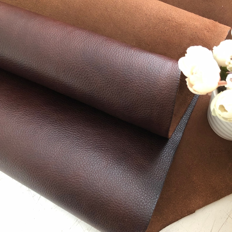 May include: Two pieces of leather, one brown and one dark brown, are folded over each other. The brown leather has a smooth texture, while the dark brown leather has a pebbled texture.