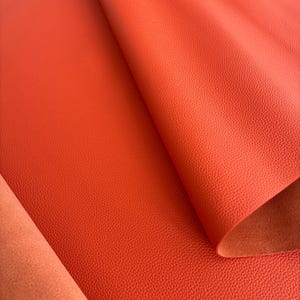 May include: Close-up of a vibrant orange leather sheet with a textured surface. The leather is folded, revealing a lighter orange underside. The material appears to be high-quality, suitable for various crafting projects.