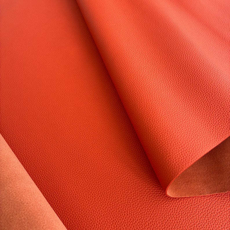 May include: Close-up of a vibrant orange leather sheet with a textured surface. The leather is folded, revealing a lighter orange underside. The material appears to be high-quality, suitable for various crafting projects.