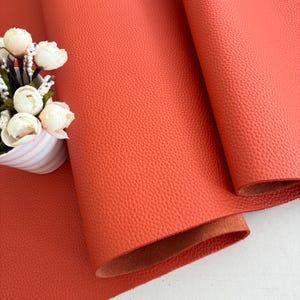 May include: Close-up of a vibrant orange leather sheet with a textured surface. The leather is folded, revealing a glimpse of the underside. A white vase with white flowers is in the background.