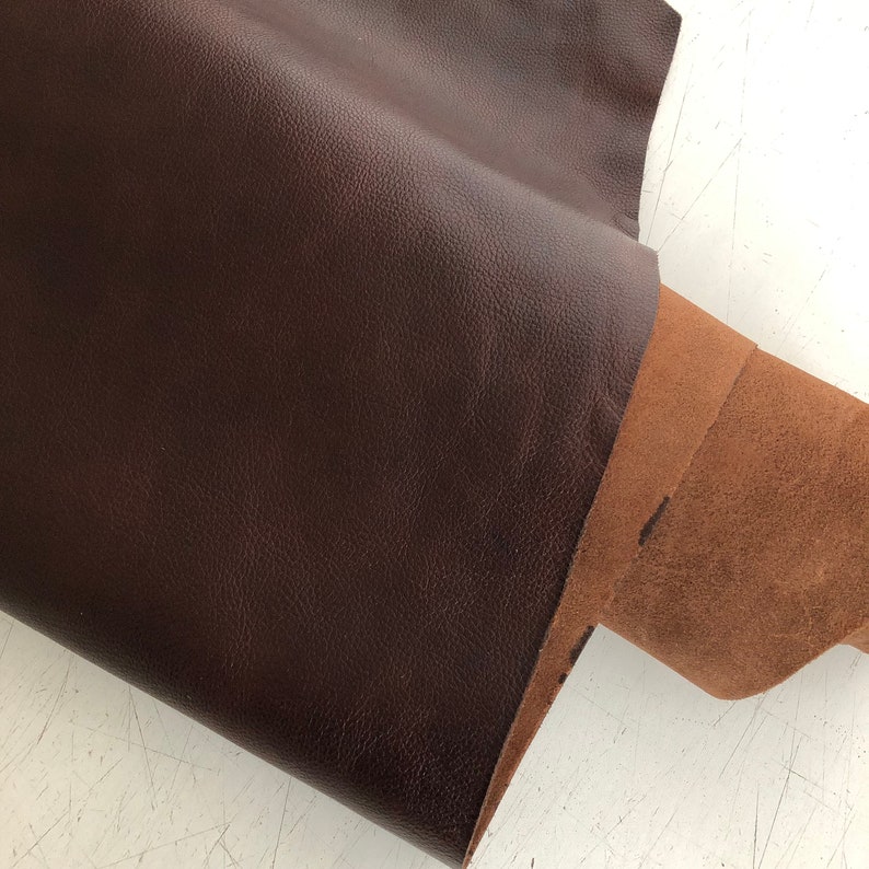 May include: A close-up of a brown leather sheet folded over a brown suede sheet. The leather has a pebbled texture.