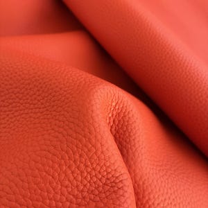 May include: Close-up of vibrant orange leather with a textured, pebbled surface. The leather is folded, revealing the grain and natural variations in the material. The color is consistent throughout, with subtle highlights and shadows.