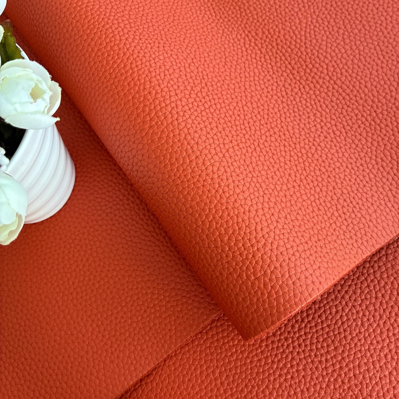 May include: Close-up of a textured, orange leather material. The leather has a pebbled grain and is folded over, revealing the underside. A white vase with white flowers is in the upper left corner.