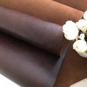 May include: Two pieces of leather fabric, one brown and one dark brown, are folded over each other. The brown fabric has a smooth texture, while the dark brown fabric has a pebbled texture.
