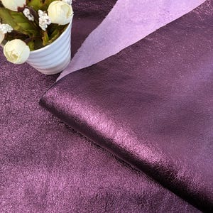 Pode incluir: Close-up de tecido roxo brilhante e texturizado. O tecido tem um brilho metálico e uma aparência ligeiramente enrugada. Um tecido roxo mais claro é sobreposto atrás do tecido principal. Um vaso de cerâmica branco com flores brancas está no canto superior esquerdo.