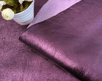 Pode incluir: Close-up de tecido roxo brilhante e texturizado. O tecido tem um brilho metálico e uma aparência ligeiramente enrugada. Um tecido roxo mais claro é sobreposto atrás do tecido principal. Um vaso de cerâmica branco com flores brancas está no canto superior esquerdo.