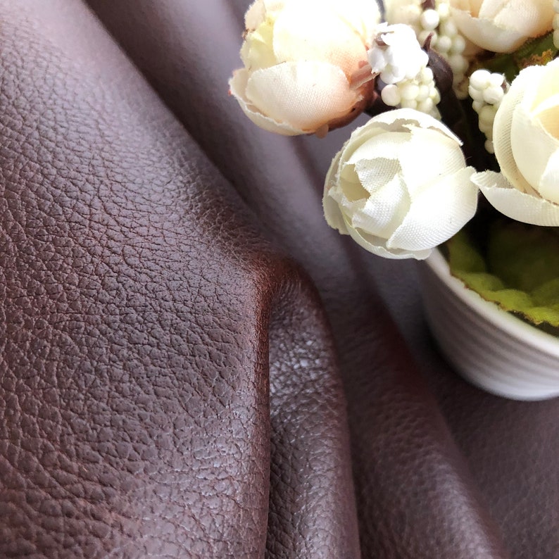 May include: Close-up of a brown leather fabric with a textured surface. A bouquet of white flowers with green leaves is in the background.