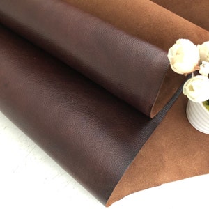May include: Two pieces of leather, one brown and one dark brown, are folded together. The brown leather has a smooth texture, while the dark brown leather has a pebbled texture. A small bouquet of white flowers is sitting on the brown leather.