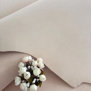 May include: A close-up of a light beige leather hide with a soft, matte finish. A small bouquet of white roses with green leaves sits in the lower left corner. The leather has a smooth texture and a natural appearance.