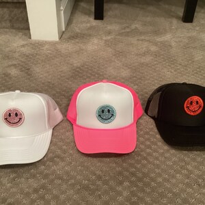 May include: Three trucker hats with different colored brims and smiley face patches. The hats are white, pink, and black. The smiley faces are pink, blue, and red.
