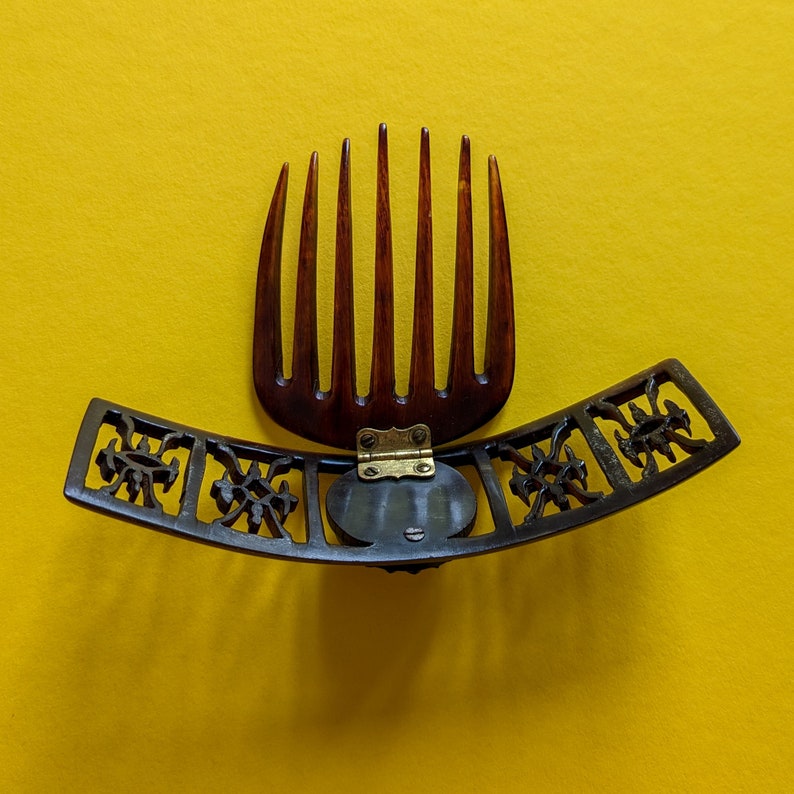 May include: A brown and black hair comb with a decorative metal hinge. The comb has a curved, ornate design with floral motifs.