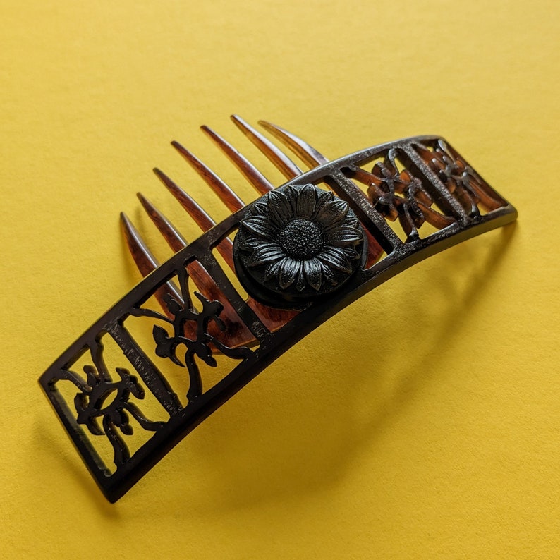 May include: A black plastic hair comb with a floral design and a black flower accent. The comb has a curved shape and is designed to hold hair in place.