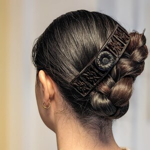 May include: A woman with dark brown hair is wearing a black hair clip with intricate floral designs. The clip is holding her hair in a braided bun.