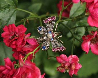 Könnte beinhalten: Eine goldene Schmetterlingsbrosche mit einer Perle, Saphiren und Rubinen. Die Brosche ist vor einem Hintergrund aus rosa Rosen platziert.
