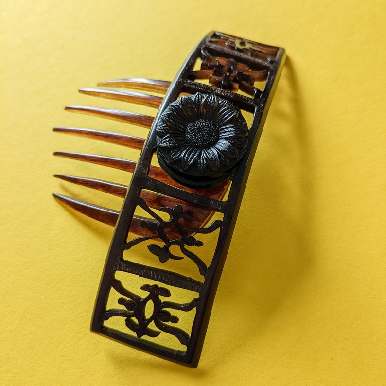 May include: A black plastic hair comb with a decorative floral design and a black flower accent. The comb has a row of teeth on one side and a decorative design on the other.