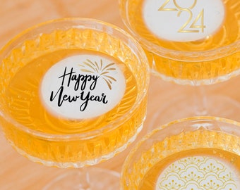 May include: Three clear glass champagne flutes with gold and black decorative stickers. The stickers say "Happy New Year", "2024", and a geometric pattern.