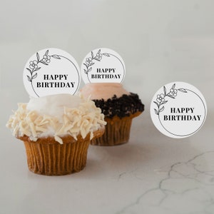 May include: Two cupcakes with white and black frosting topped with white chocolate curls. Each cupcake has a round white paper topper with a black floral design and the text "Happy Birthday".