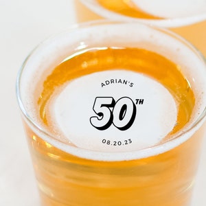 May include: A glass of beer with a white sticker that says "Adrian's 50th 08.20.23"