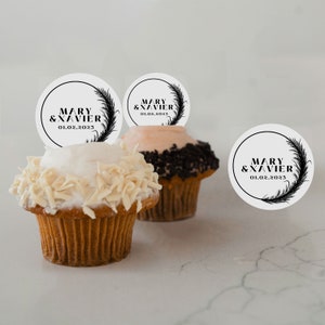 May include: Cupcakes with white and chocolate frosting, topped with decorative toppers. The toppers are white circles with the text "MARY & XAVIER 01.02.2023" in black, surrounded by a feather design.