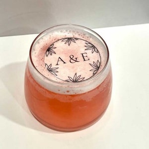 Pode incluir: Um copo transparente com uma bebida rosa e um adesivo branco com desenhos florais pretos e o texto "A&E" no topo.