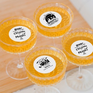 May include: Four clear glass champagne flutes with white round stickers. The stickers have black text and illustrations. The text on the stickers reads "Happy Christmas Muggles", "I'm Dreaming of a Hogwarts Christmas", and "It's the Most Magical Time of the Year".