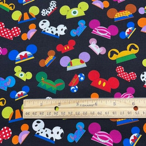 May include: Black fabric featuring a repeating pattern of Mickey Mouse ears in various colors and designs. The ears include polka dots, stripes, and character-themed styles. A ruler is placed across the fabric, showing measurements in inches.