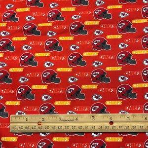 May include: Red fabric with a repeating pattern of white football helmets with the word "Chiefs" in yellow. The fabric is measured with a wooden ruler.