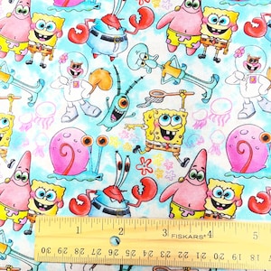 May include: Fabric featuring a repeating pattern of cartoon characters, including a yellow sponge, a pink starfish, and a blue crab, set against a light blue background. The design includes a ruler with inch measurements.