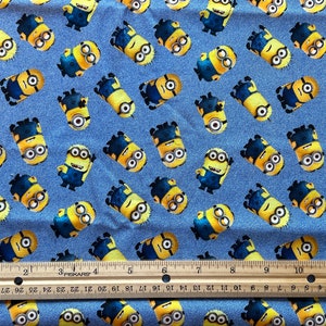 May include: Blue denim fabric with a repeating pattern of yellow Minions. The Minions are wearing blue overalls and goggles. A wooden ruler is in the bottom of the image.