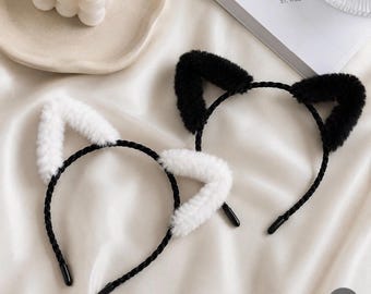 May include: Two cat ear headbands. One headband has black ears, and the other has white ears. The ears are fluffy and attached to a black headband. The headbands are on a white satin surface.