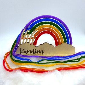 Op de afbeelding: Houten regenboog-ambacht met een wolkenbasis en de naam "Karolina" erop gegraveerd. De regenboog is gemaakt van kleurrijk garen in rood, oranje, geel, groen, blauw en paars.