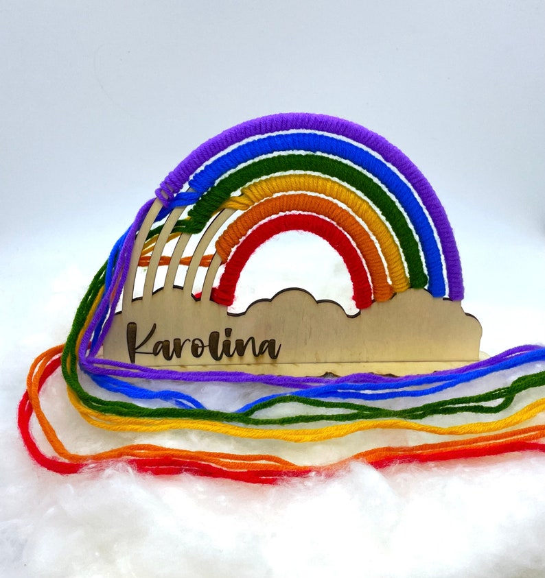 Op de afbeelding: Houten regenboog-ambacht met een wolkenbasis en de naam "Karolina" erop gegraveerd. De regenboog is gemaakt van kleurrijk garen in rood, oranje, geel, groen, blauw en paars.