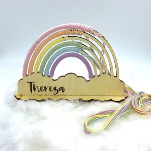 Op de afbeelding: Een houten regenboog met pastelkleurig garen eromheen gewikkeld. De regenboog is bevestigd aan een houten wolk met de naam "Theresa" erop gegraveerd.