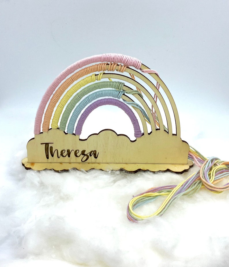 Op de afbeelding: Een houten regenboog met pastelkleurig garen eromheen gewikkeld. De regenboog is bevestigd aan een houten wolk met de naam "Theresa" erop gegraveerd.