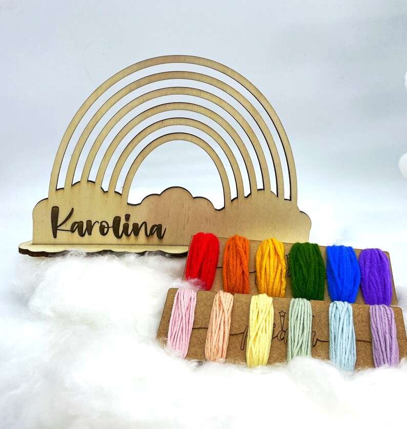 Op de afbeelding: Houten regenboog garen houder met de naam "Karolina" gegraveerd op de basis. De houder wordt geleverd met een set kleurrijke garenklossen in regenboogkleuren.