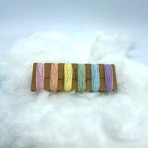 Op de afbeelding: Een set van zes pastelkleurige borduurgaren spoelen op een houten kaart. De kleuren zijn roze, perzik, geel, groen, blauw en lavendel.