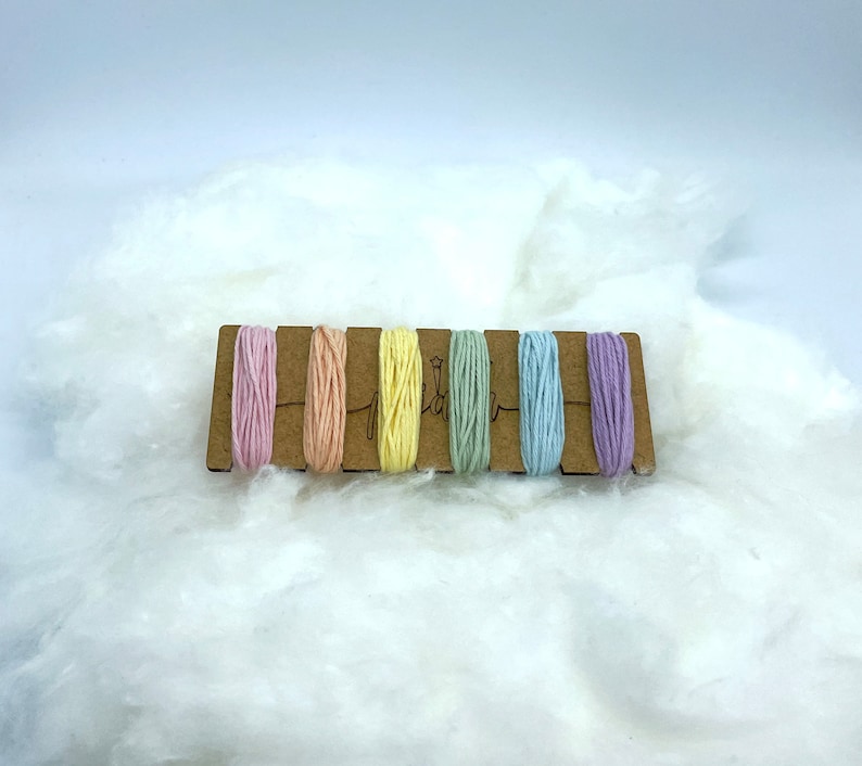 Op de afbeelding: Een set van zes pastelkleurige borduurgaren spoelen op een houten kaart. De kleuren zijn roze, perzik, geel, groen, blauw en lavendel.