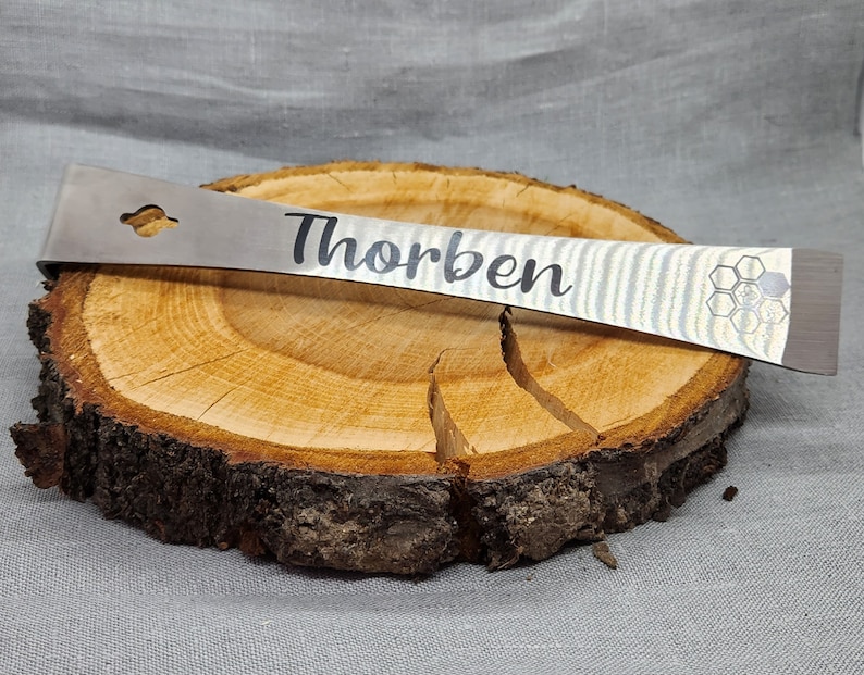 Puede incluir: Una palanca de metal plateada con un patr&oacute;n de panal y el nombre "Thorben" grabado en ella. La palanca est&aacute; apoyada en un tronco de madera.