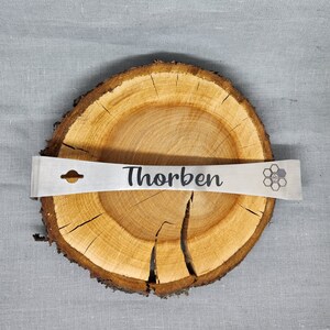 Puede incluir: Una barra de palanca de metal plateado con un agujero en el mango y la palabra "Thorben" grabada en ella. La barra de palanca est&aacute; apoyada en una rodaja de madera con un dise&ntilde;o de panal en el lado derecho.