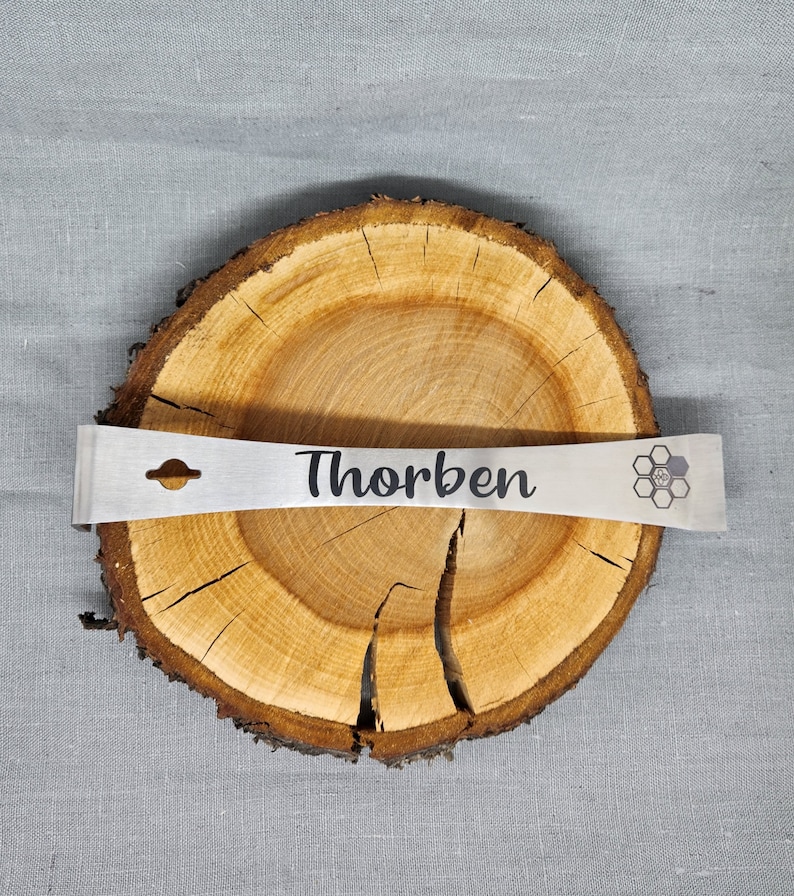 Puede incluir: Una barra de palanca de metal plateado con un agujero en el mango y la palabra "Thorben" grabada en ella. La barra de palanca est&aacute; apoyada en una rodaja de madera con un dise&ntilde;o de panal en el lado derecho.