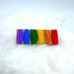 Op de afbeelding: Een set van zes borduurgaren spoelen in regenboogkleuren, waaronder paars, blauw, groen, geel, oranje en rood, gerangschikt op een houten kaart.
