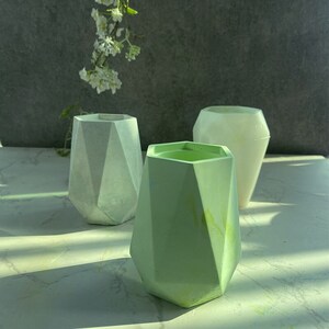 May include: Three geometric concrete planters in different shades of green. The planters are all in the shape of a hexagon with rounded edges.