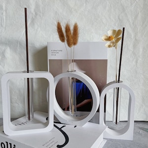 Può includere: Tre vasi geometrici bianchi con provette di vetro trasparente per contenere fiori secchi e un singolo stelo. I vasi sono disposti in fila su una superficie bianca. Il testo "Furniture Design Idea" è visibile su una rivista sullo sfondo.