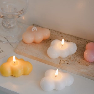 May include: Four cloud-shaped candles in yellow, peach, white, and pink. The candles are lit and have a soft glow.