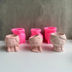 May include: Four small pink and beige ceramic planters shaped like girls with closed eyes. Three of the planters are plain pink and one is a beige girl with a braid.