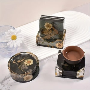 May include: Set of four black and white coasters with pressed flowers and leaves. The coasters are stacked and displayed on a white surface. A brown ceramic cup is sitting on top of the coasters.